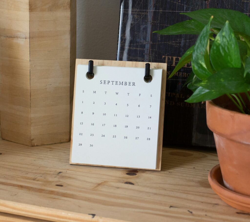 Desktop calendar showing September dates, placed on a wooden shelf beside a green potted plant and books.