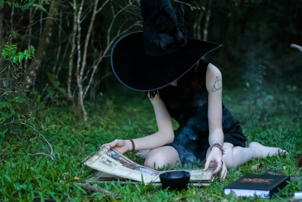 Witch in forest reading spellbook, wearing a hat with feather detail, surrounded by magical objects on grass.