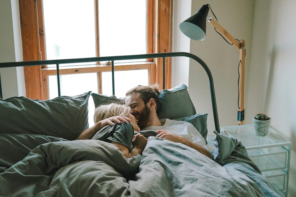 Couple cuddling in bed under grey duvet, next to window with natural light, creating a cosy, intimate atmosphere.