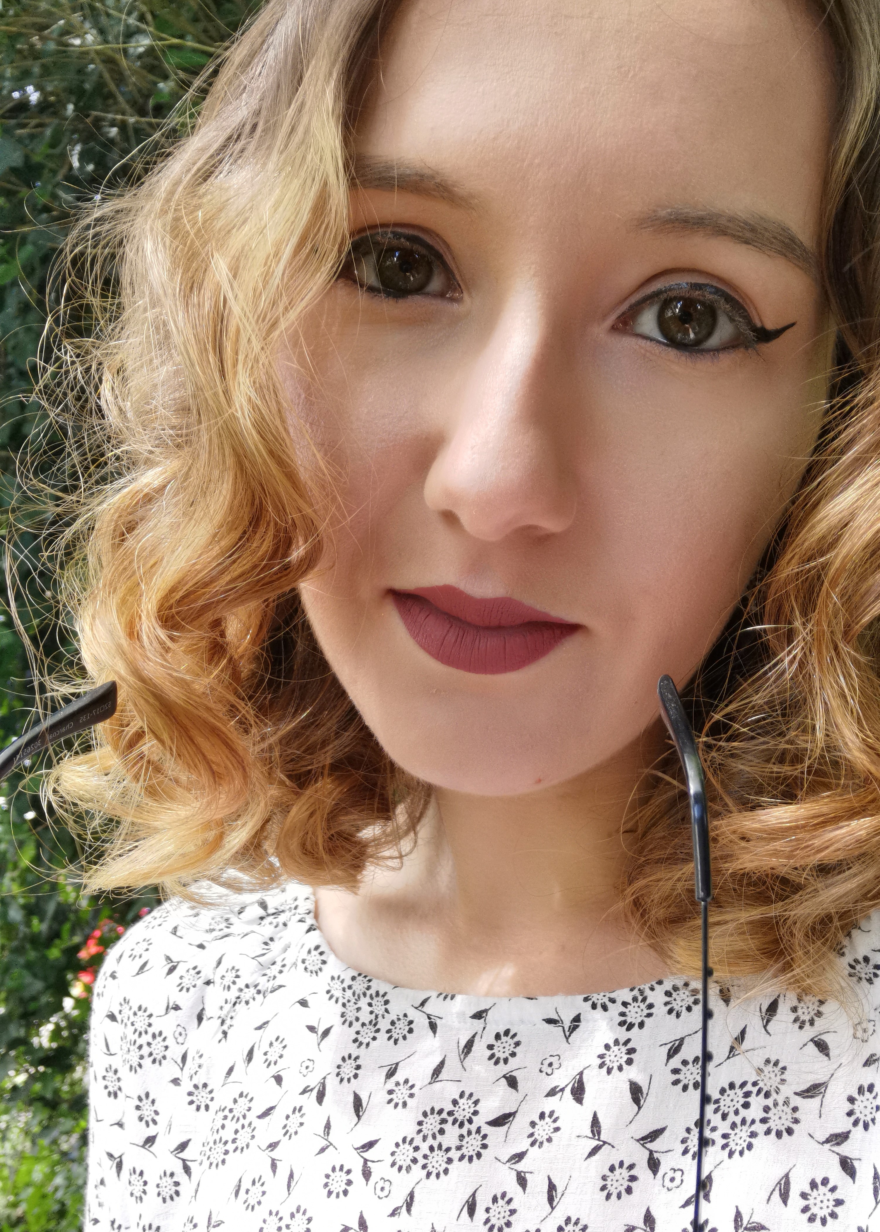 Young woman with curly hair and floral dress poses outdoors, vibrant makeup and a soft smile.