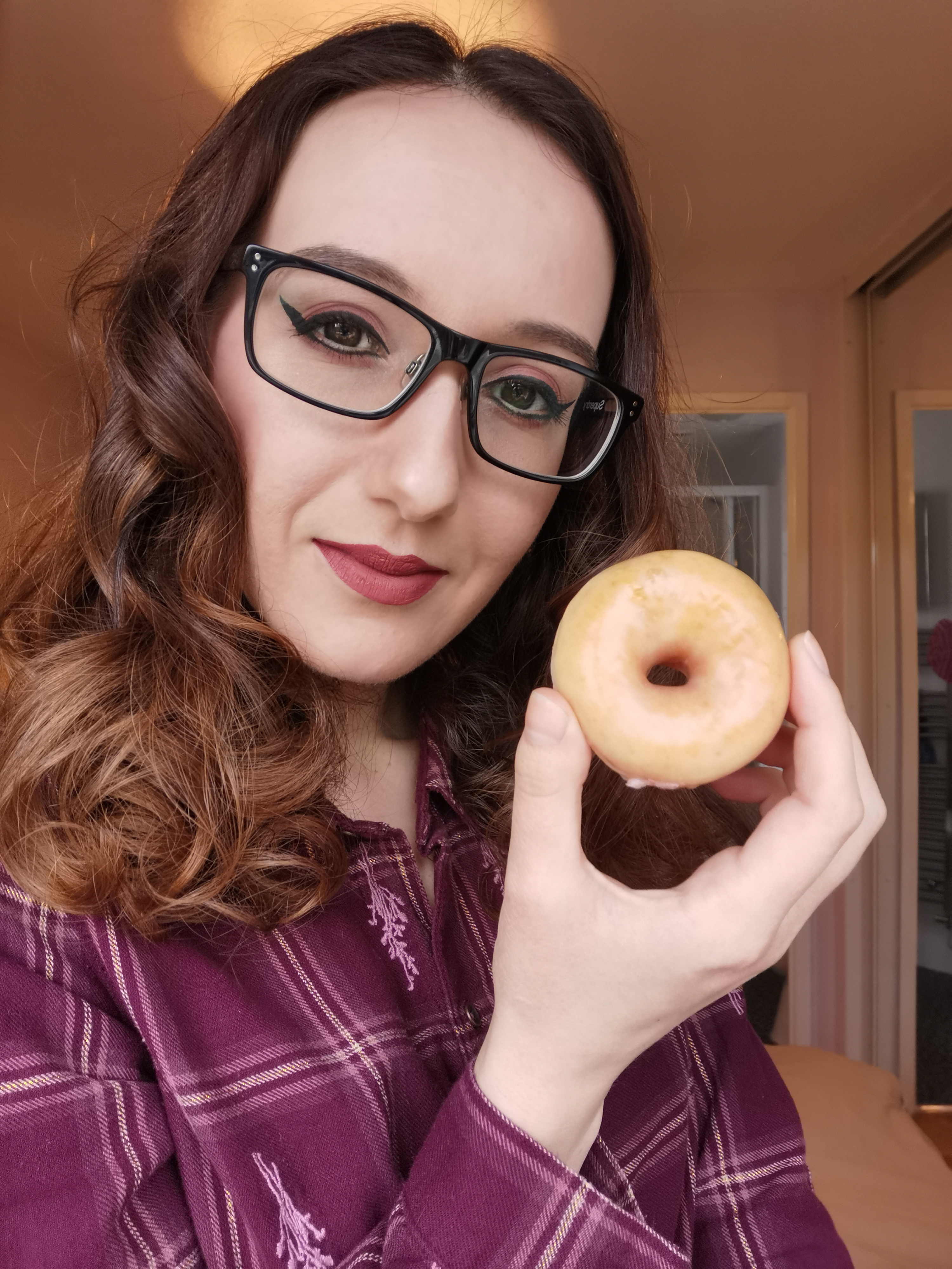 Person with glasses holding a glazed doughnut, wearing a purple checkered shirt, smiling indoors.