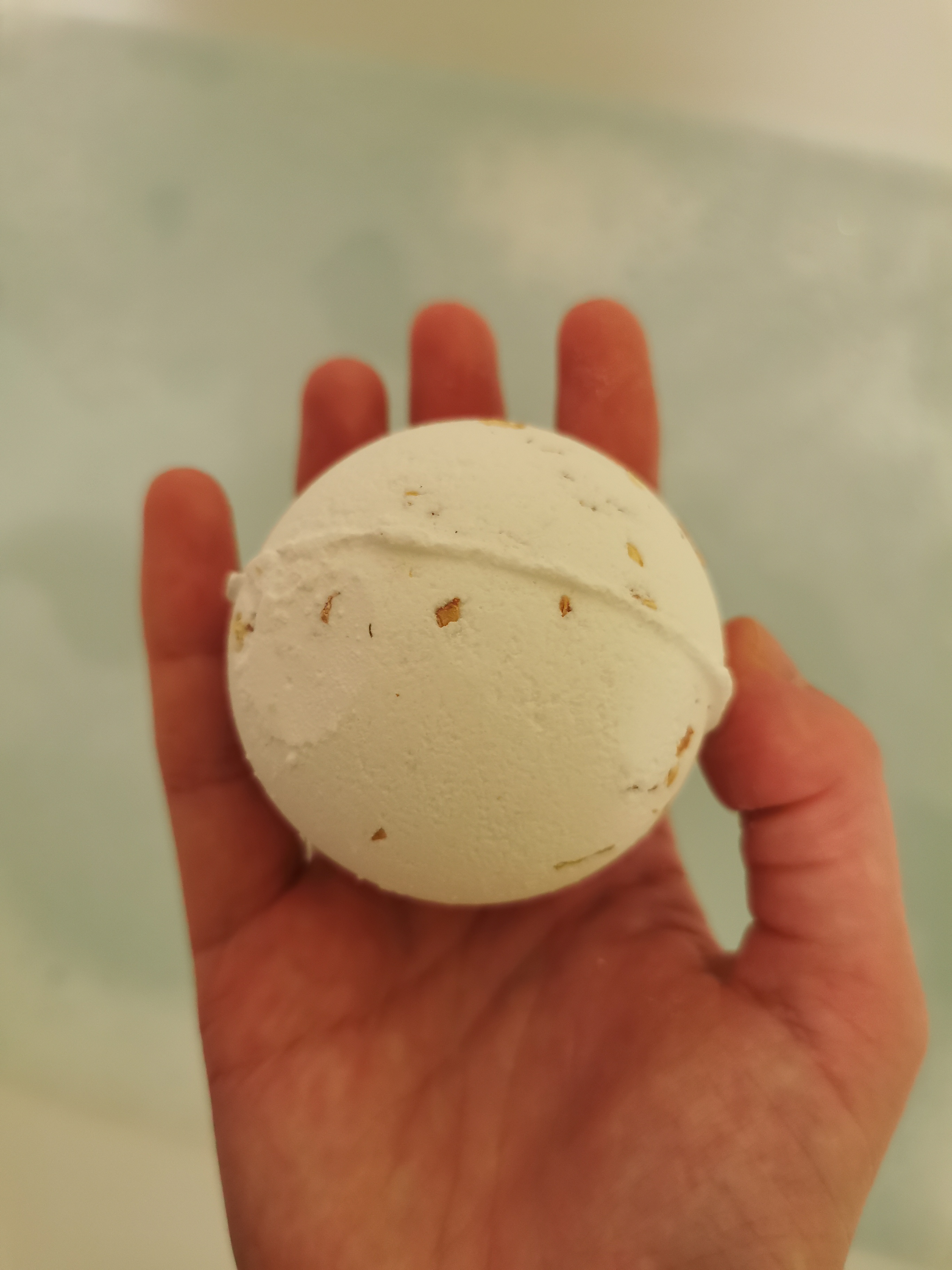 Hand holding a white bath bomb with speckled texture above bath water, ready to create a relaxing spa experience.