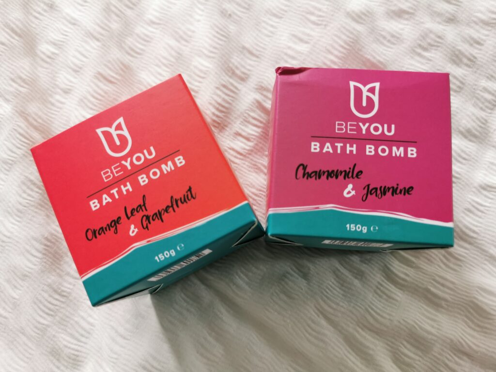 Two BeYou bath bomb boxes: Orange Leaf & Grapefruit and Chamomile & Jasmine, 150g each, on a textured surface.