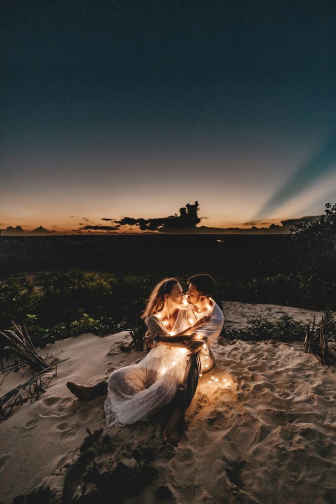 Romantic couple wrapped in fairy lights on a sandy beach at sunset, with a serene sky in the background.