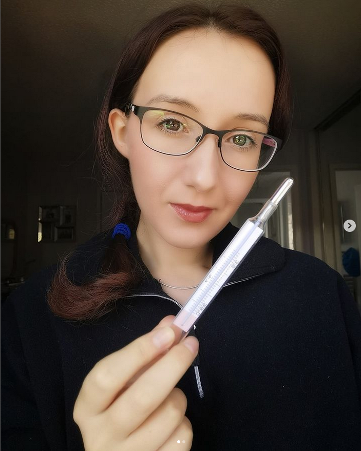 Person with glasses holding a thermometer, wearing a black top, indoors with soft lighting.