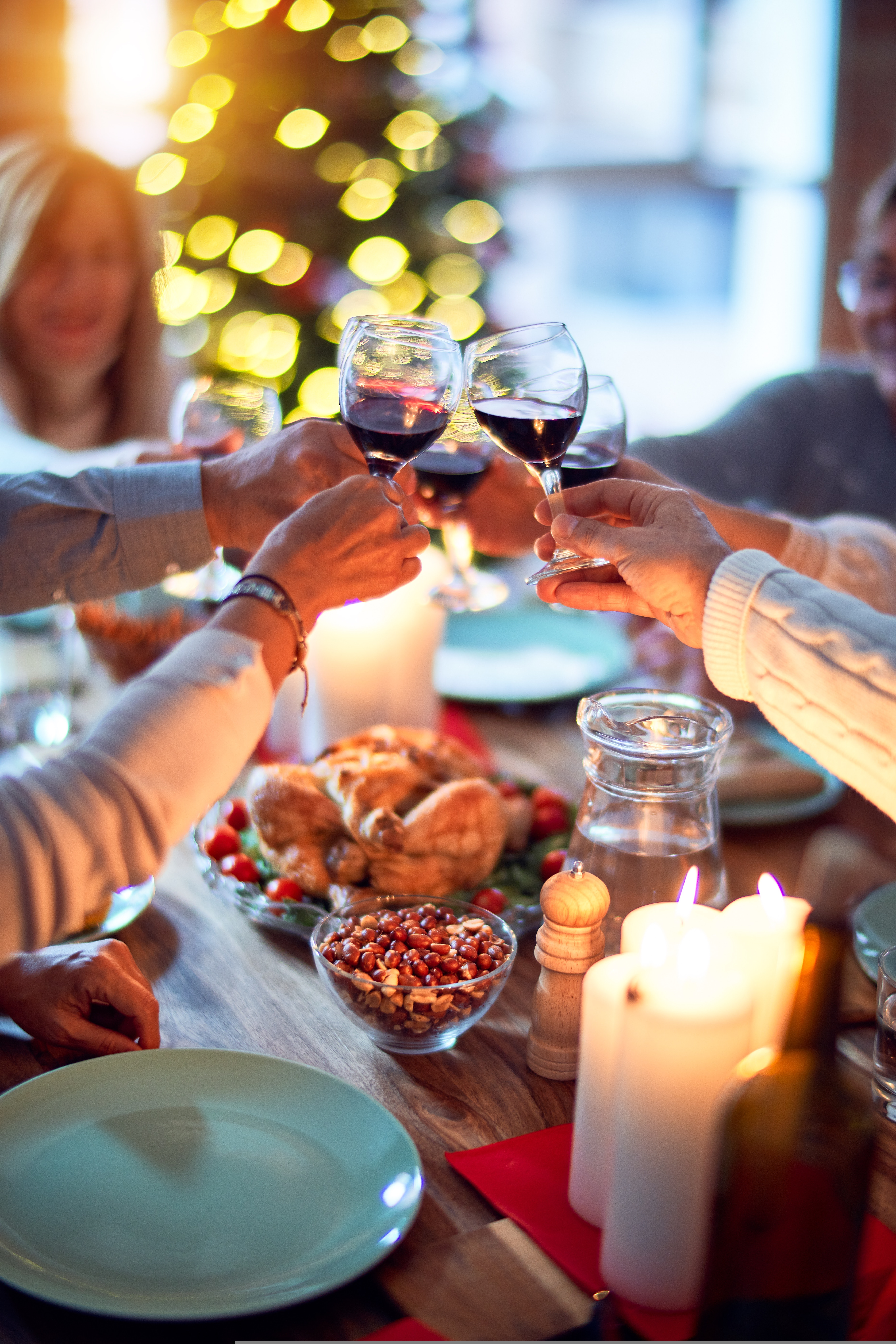 Festive dinner with friends clinking wine glasses by candlelight, with a lit Christmas tree in the background.
