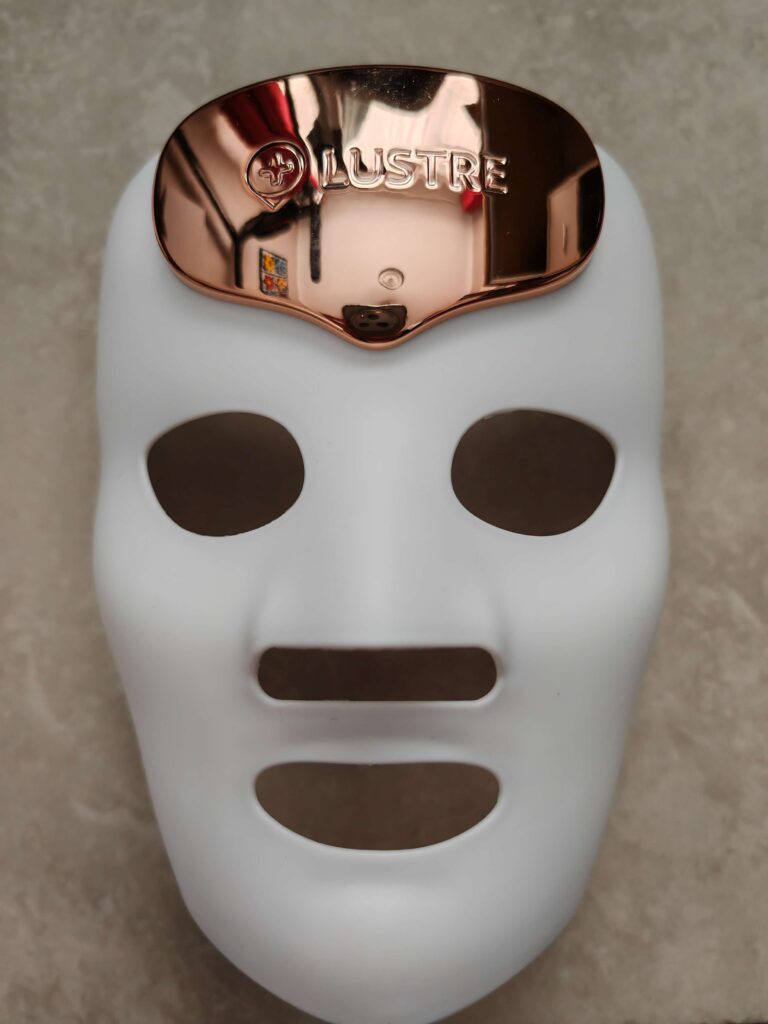 White LED facial mask with copper top by Lustre for skincare treatment on a grey background.