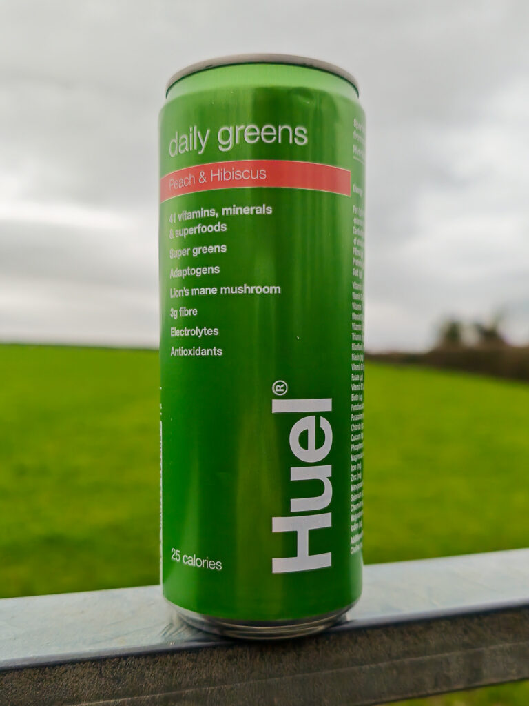 Green Huel can of daily greens with peach and hibiscus, rich in vitamins, fibre, and superfoods, in a field setting.