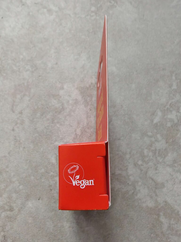 orange packaging with 'Vegan' label and sunflower icon on a grey surface, highlighting plant-based product branding.