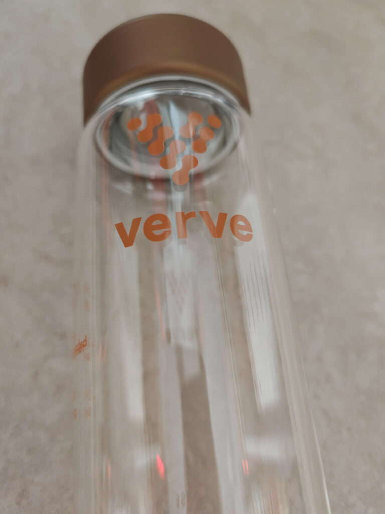 Clear glass water bottle with brown lid and verve text, featuring orange logo, resting on a light surface.