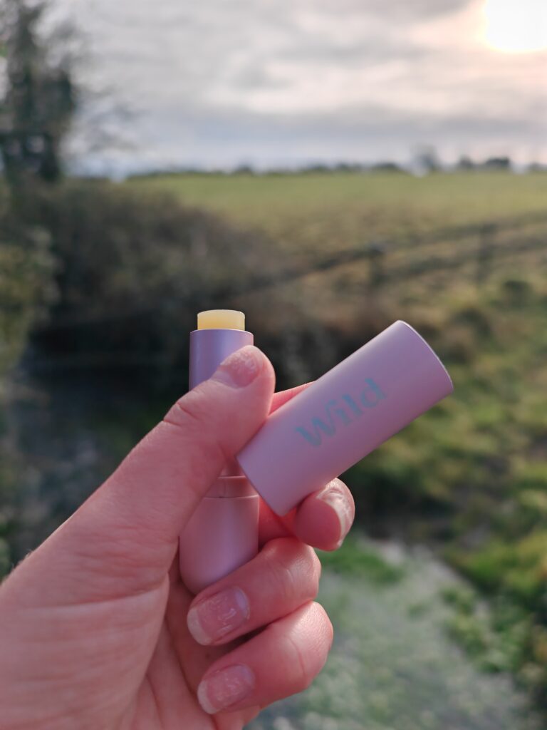 Hand holding a pink lip balm tube outdoors with lush field in background, branded with Wild.