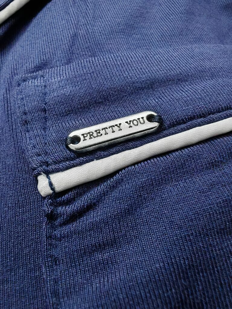Close-up of a blue fabric with a metal tag that reads Pretty You, showing stitching and design detail.