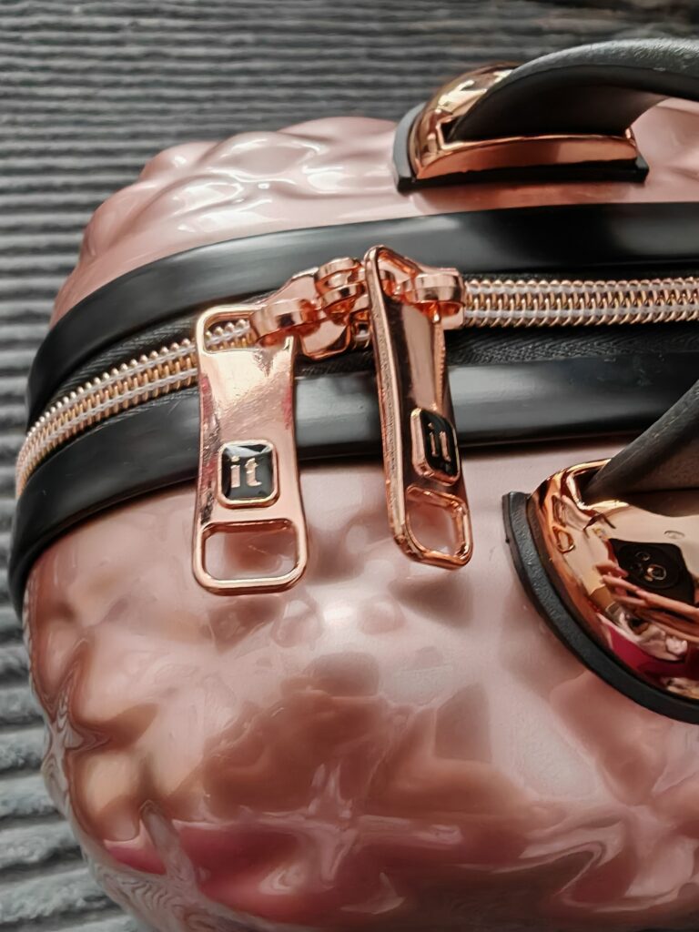 Close-up of a stylish blush pink suitcase with shiny rose gold zippers and black handles on a textured surface.