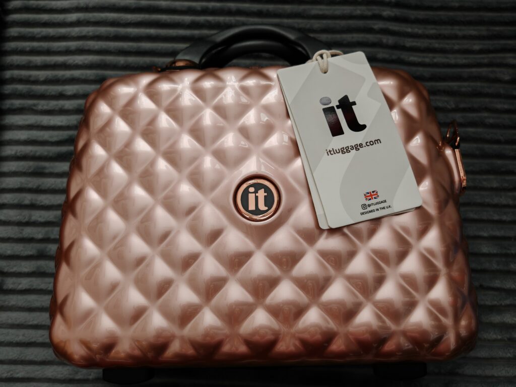 Rose gold textured suitcase by it Luggage with tag, showcasing quilted design and brand logo on gray background.