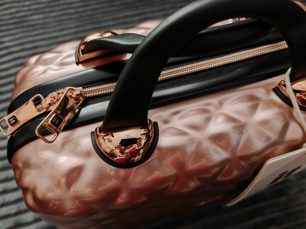 Rose gold suitcase with textured design and black handles on a striped surface, showcasing shiny zippers and accents.