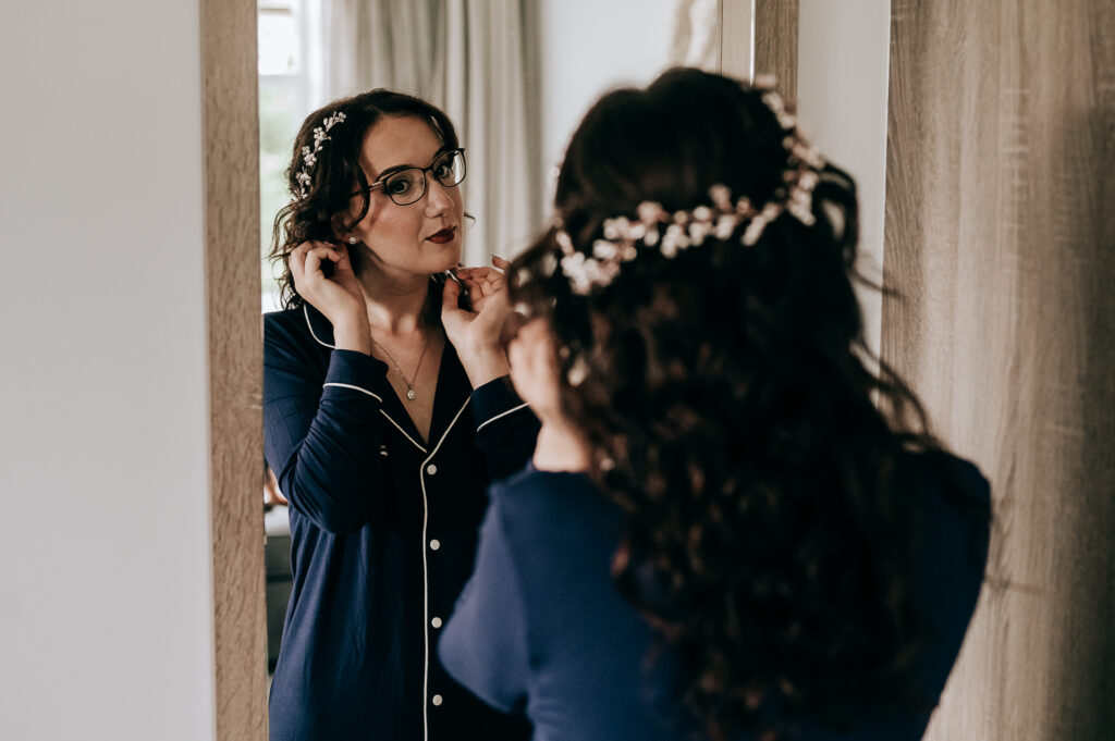 Woman in blue pajamas adjusts earrings in mirror with floral hair accessory.
