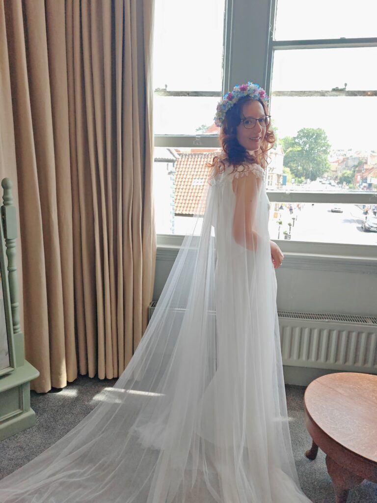 Bride in a white dress with a floral crown stands by a window, sunlight streaming in, creating a serene ambiance.