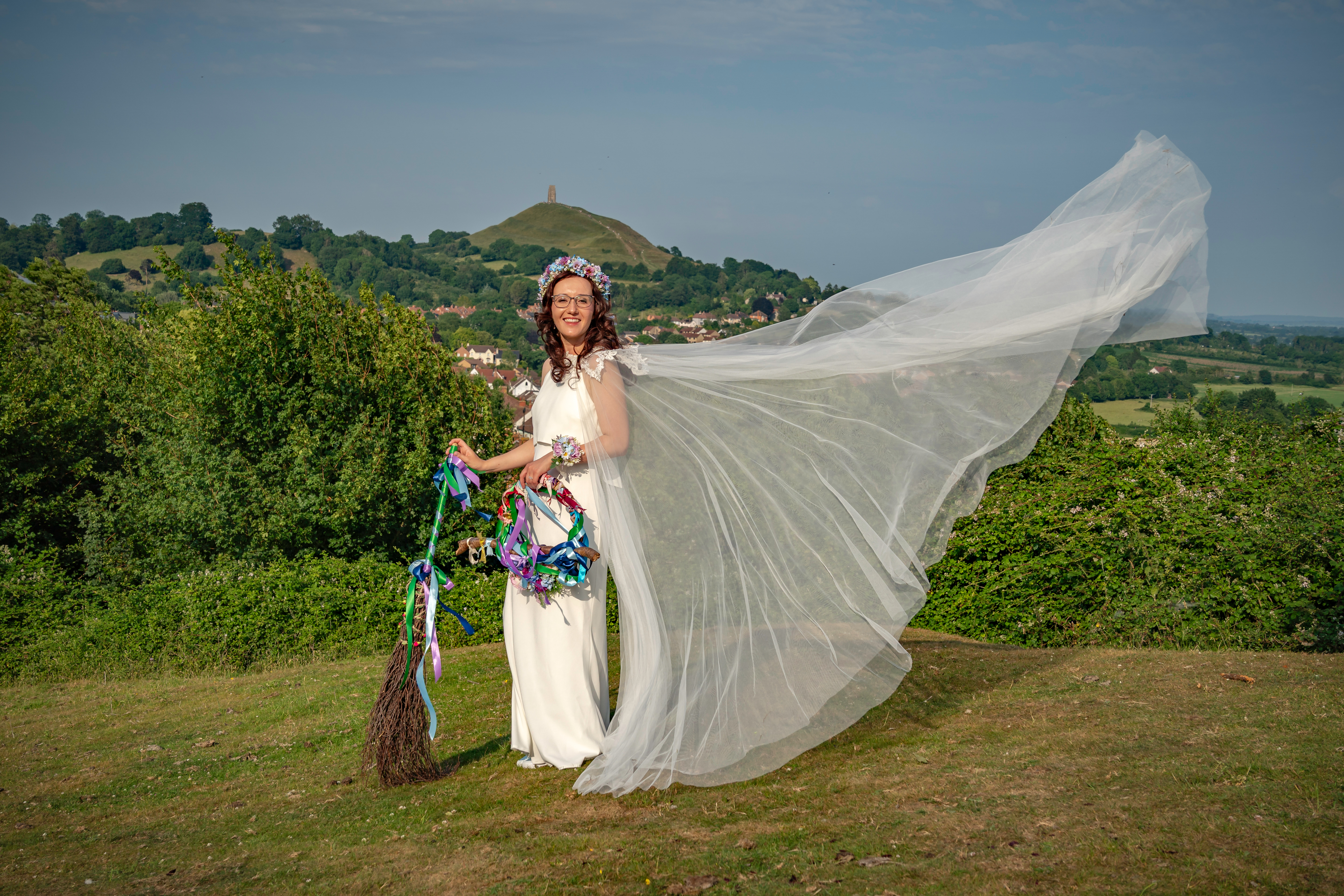 Bride in flowing gown with broom, standing on a hill with a scenic countryside backdrop and distant tower.