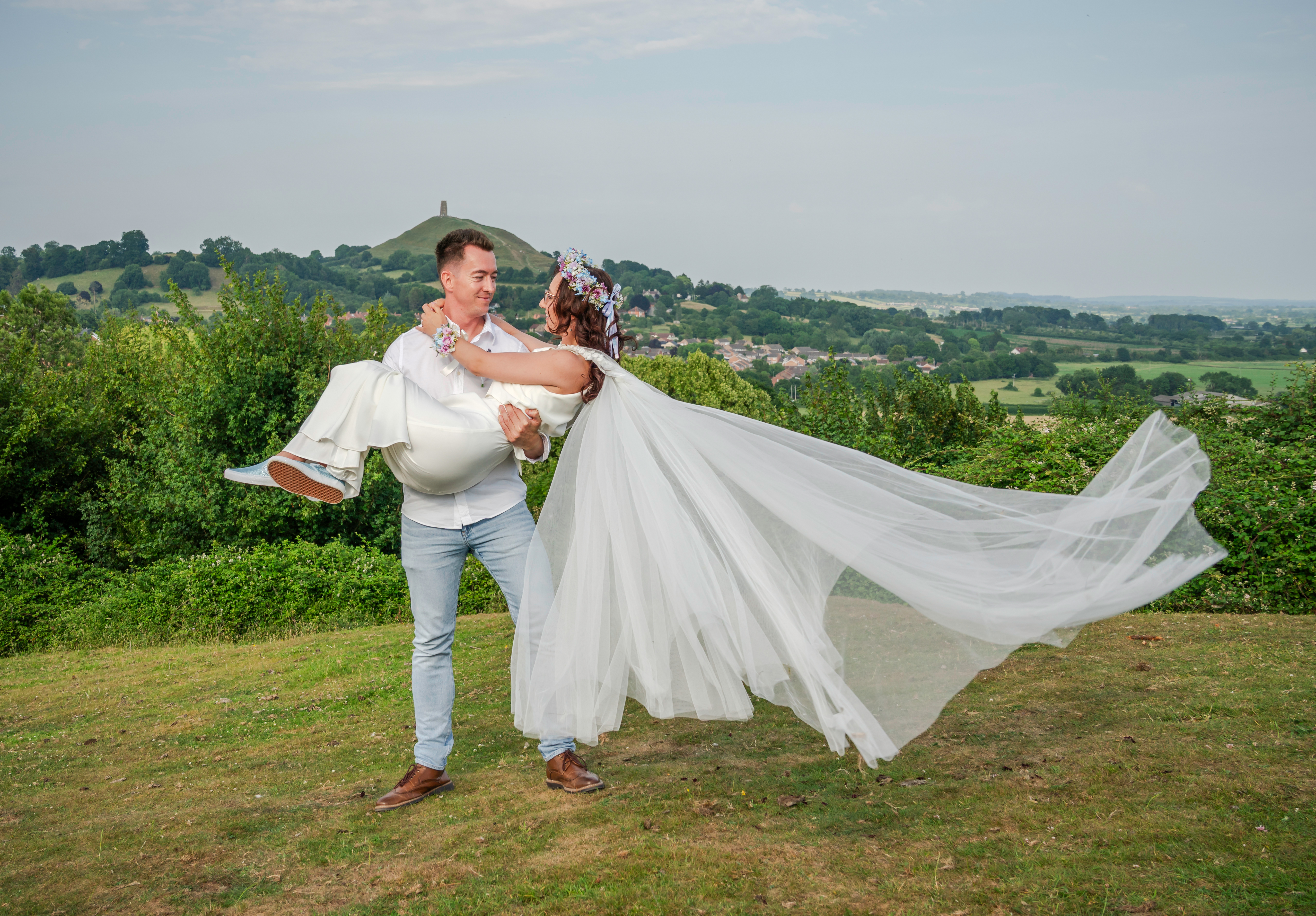 Couple joyfully celebrating wedding with scenic countryside view in background, bride being lifted by groom.