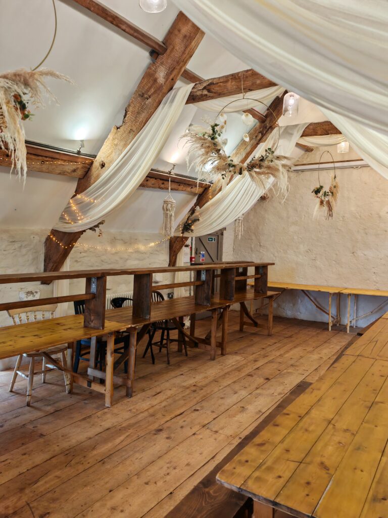 Rustic dining area with exposed wooden beams, draped fabric, and hanging greenery, set in a cozy, barn-like atmosphere.
