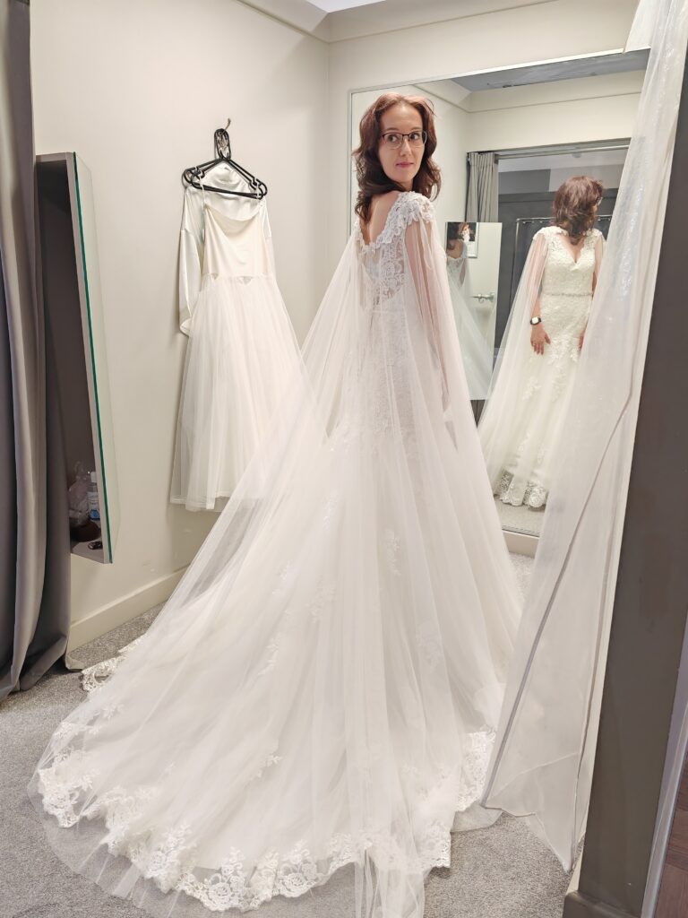 Bride trying on an elegant lace wedding dress with long train in fitting room, looking in mirror.
