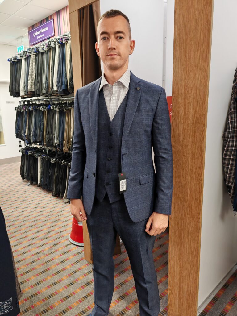 Man in a blue suit standing in a clothing store, surrounded by racks of pants.