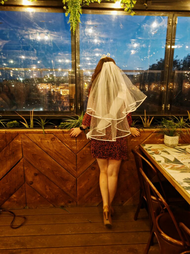 Bride-to-be in veil by a window, enjoying evening city lights at a cozy restaurant.