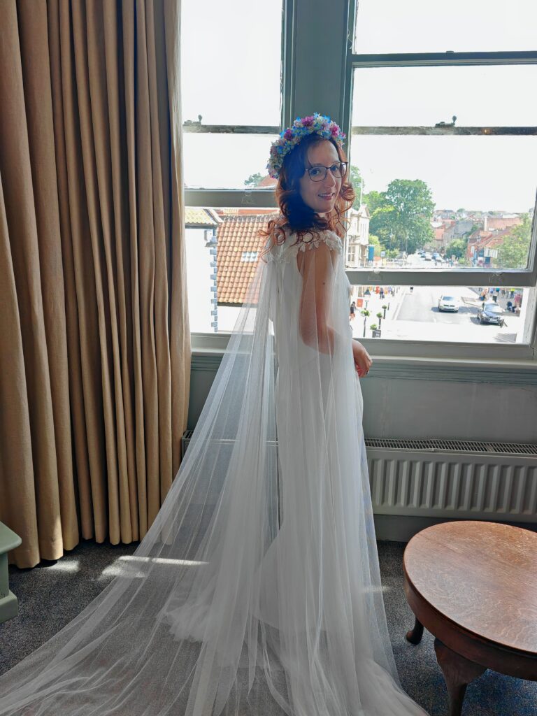 Bride in white dress and floral crown looks out a window over a picturesque street, with elegant curtains in view.