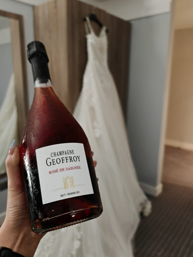 Champagne bottle in focus with an elegant white wedding dress hanging in the background.