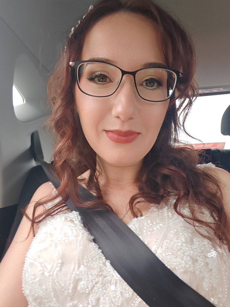 Woman with glasses and wavy hair sitting in a car wearing a beaded dress, smiling confidently at the camera.