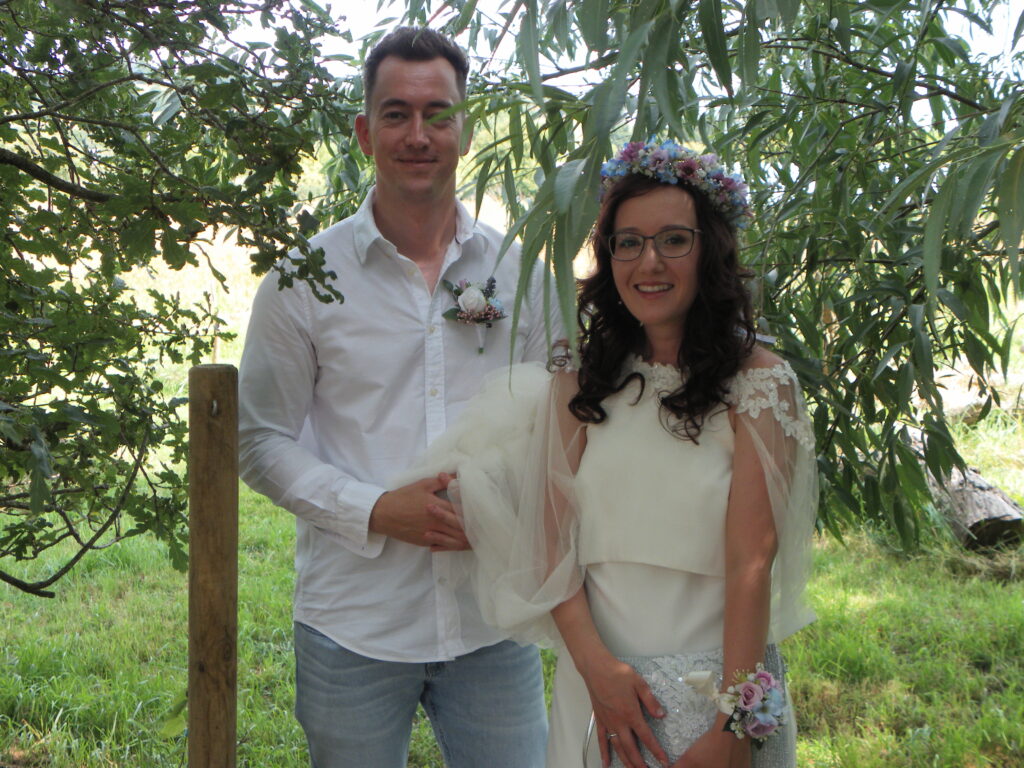 Couple in wedding attire smiles under leafy trees in outdoor setting.