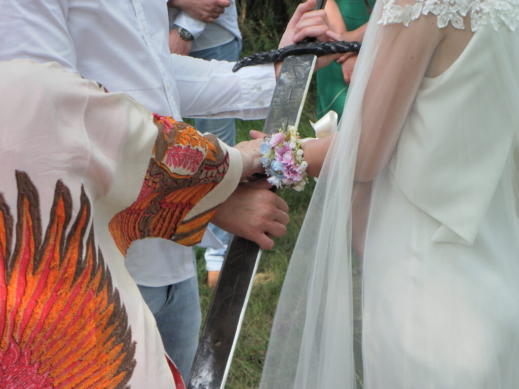 Bride and groom hold sword for a wedding ceremony, with floral dress and bouquet, symbolizing unity and tradition.