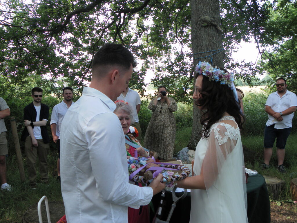 Outdoor wedding ceremony with couple and guests under trees, exchanging vows. Bride wears floral crown, groom in white shirt.