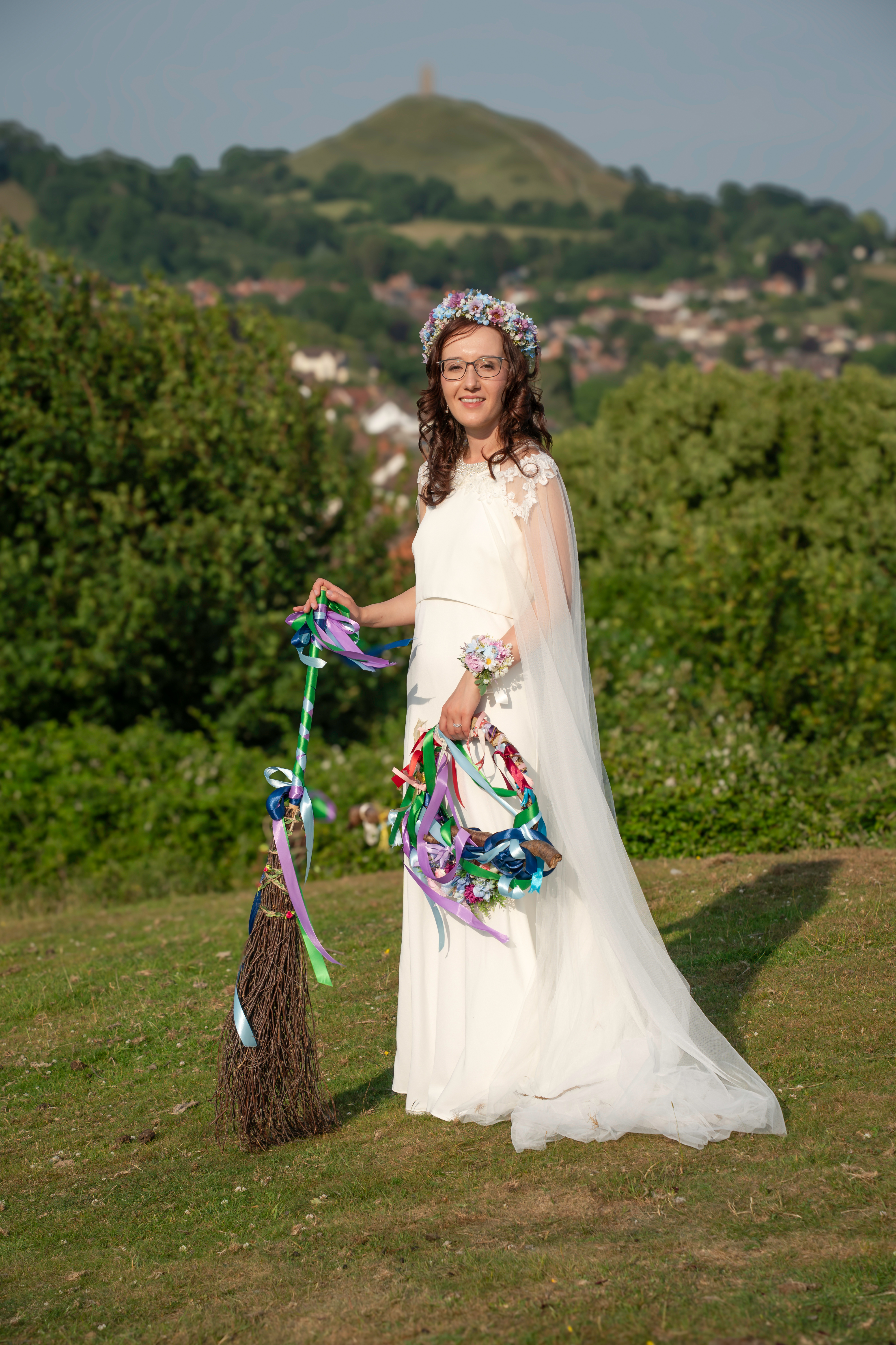 Bride in white dress with flower crown, holding broom and ribbons, outdoors near a hill.