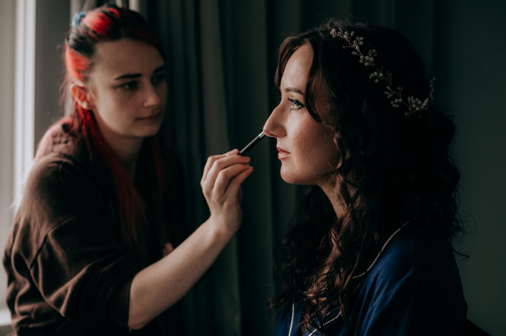 Makeup artist applying makeup on a woman in a blue robe, enhancing bridal beauty with precision and care.