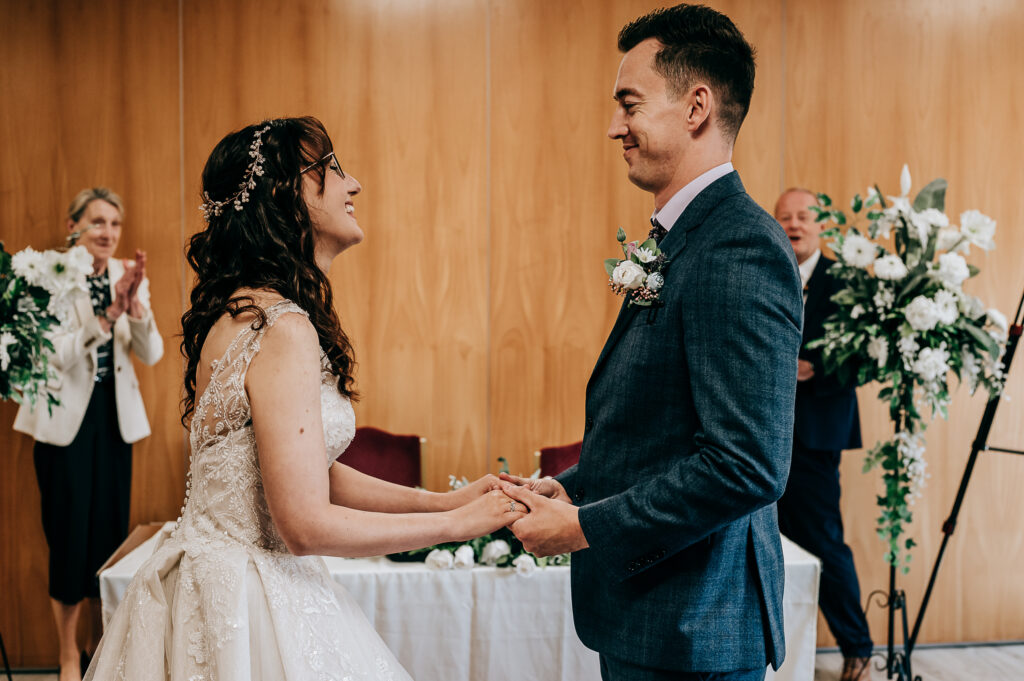 Bride and groom holding hands, smiling during wedding ceremony with guests clapping in background.