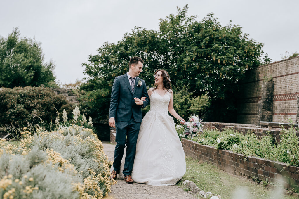 Bride and groom walking in garden, wedding dress and suit, smiling, greenery background, joyful moment.