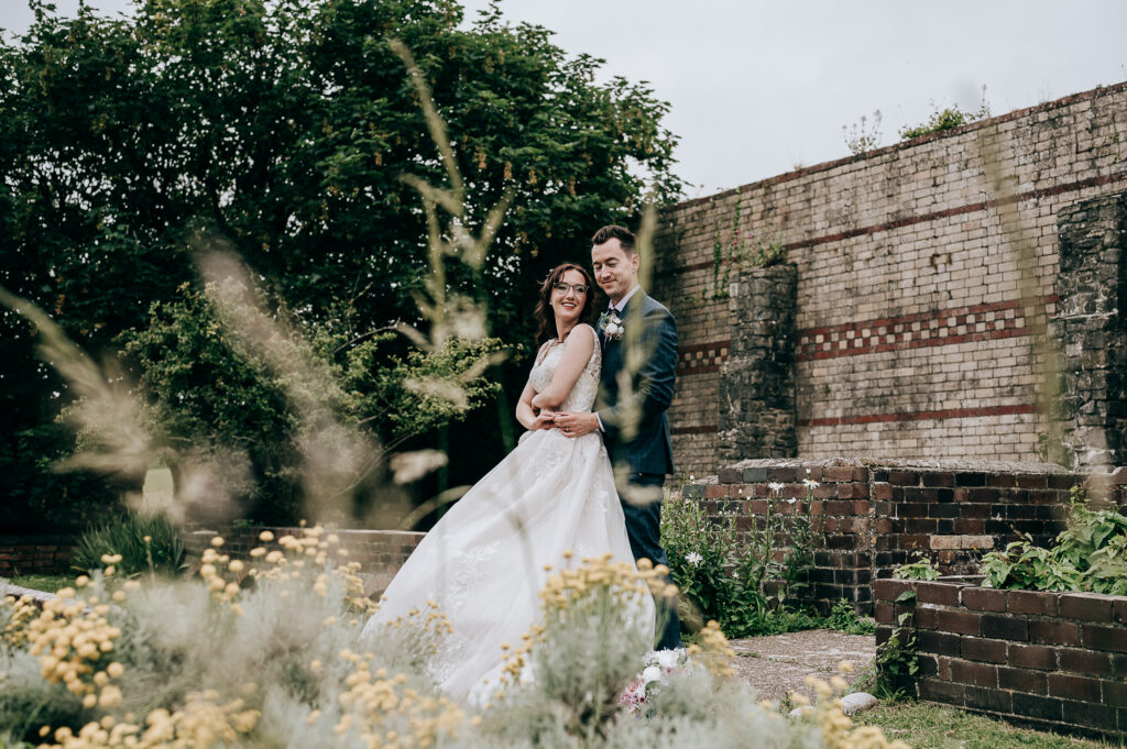Bride and groom in a garden embrace, with rustic brick wall and lush greenery in the background.