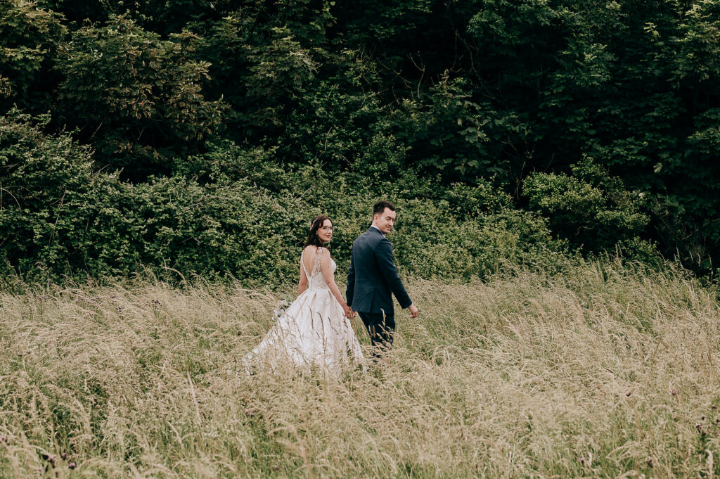 Wedding couple walking hand in hand through a grassy field with lush greenery in the background.