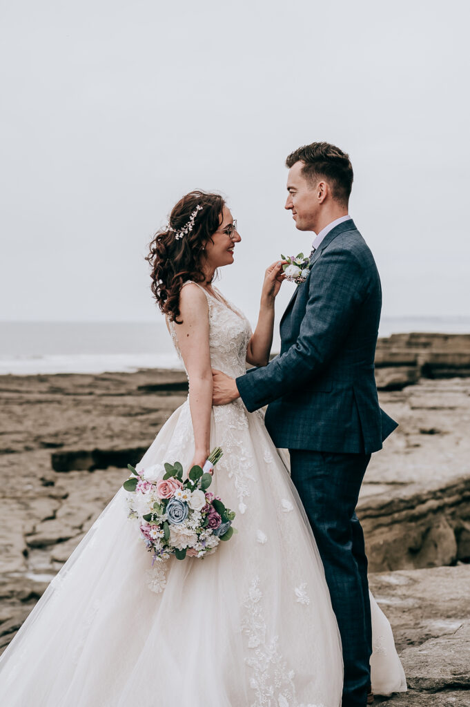 Bride and groom standing on rocky beach, holding hands and smiling, bridal bouquet in hand.