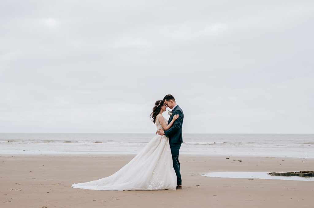 Bride and groom embrace on a serene beach, with overcast sky and ocean backdrop, capturing a romantic wedding moment.