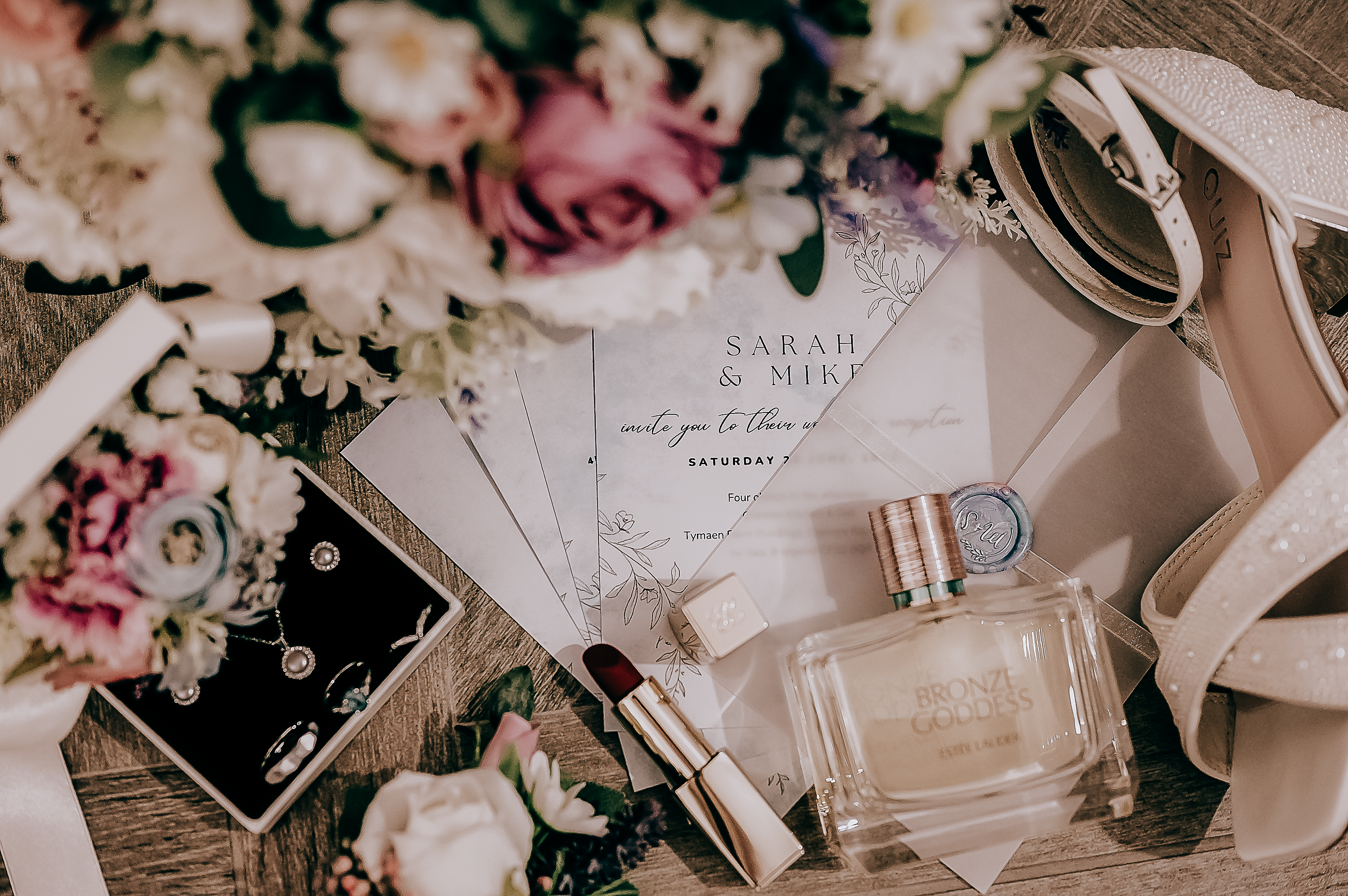 Flat lay of wedding invitation with flowers, jewelry, heels, perfume, and lipstick, creating an elegant bridal theme.