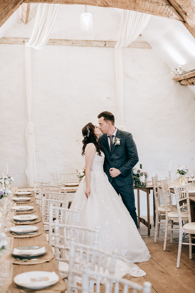 Bride and groom share a kiss in a rustic wedding venue with elegant decor and wooden tables.