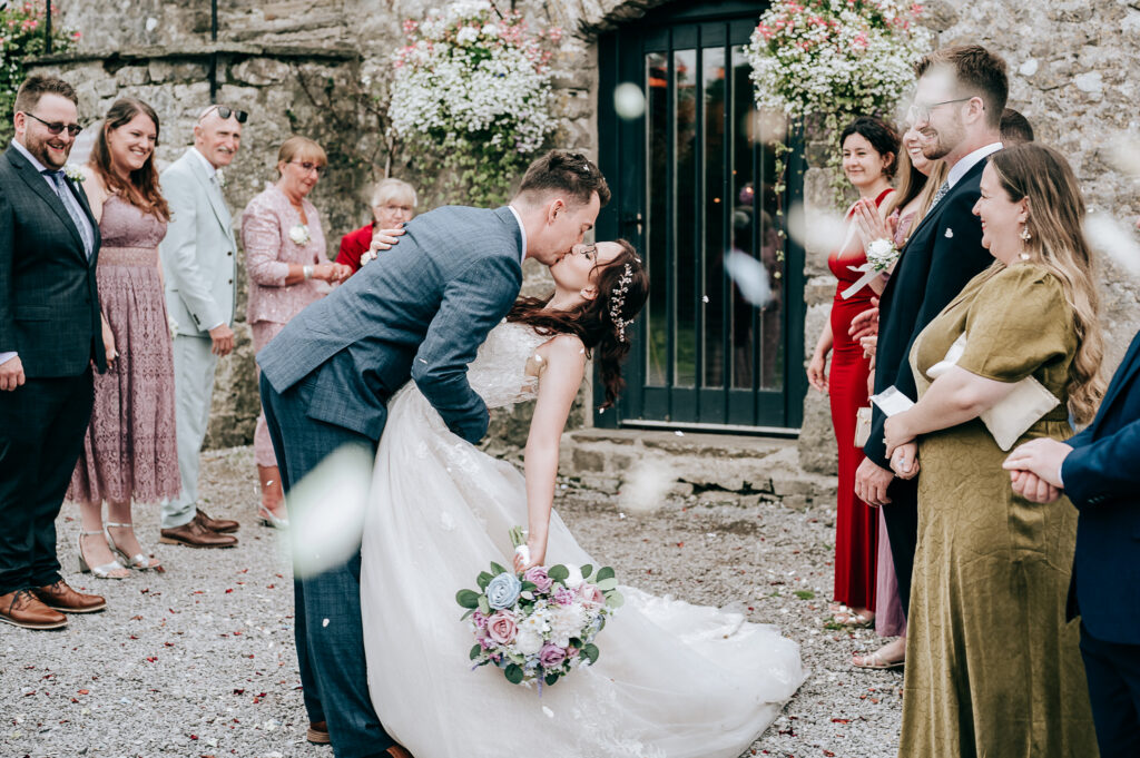 Bride and groom kiss in front of wedding guests, surrounded by floral arrangements and falling petals.