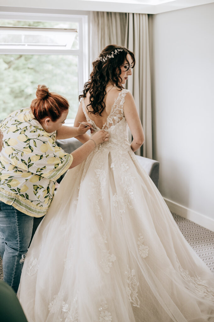 Bride getting her wedding dress adjusted by an assistant in a bright room, capturing a special pre-ceremony moment.