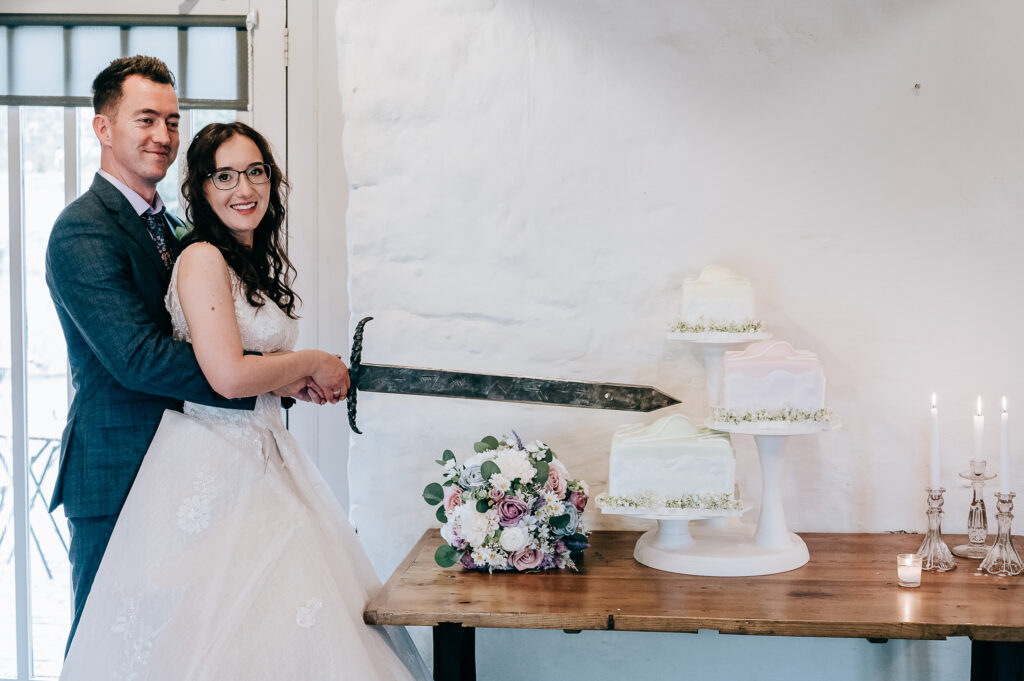 Bride and groom cutting wedding cake with sword, floral bouquet on table, elegant decor.
