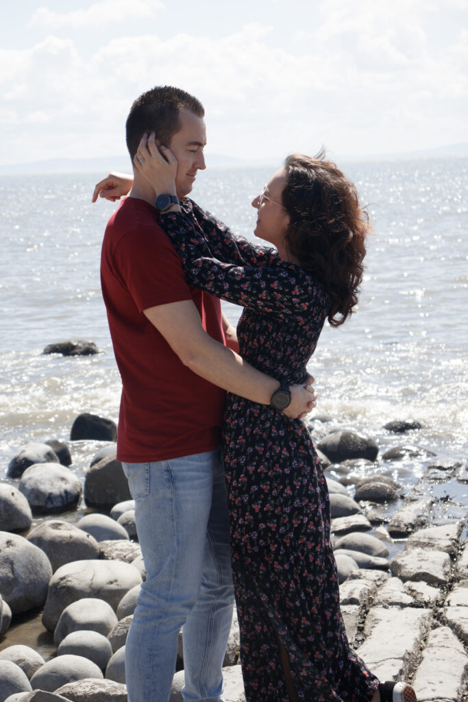 Couple embracing on rocky beach with ocean background, enjoying a sunny day together.
