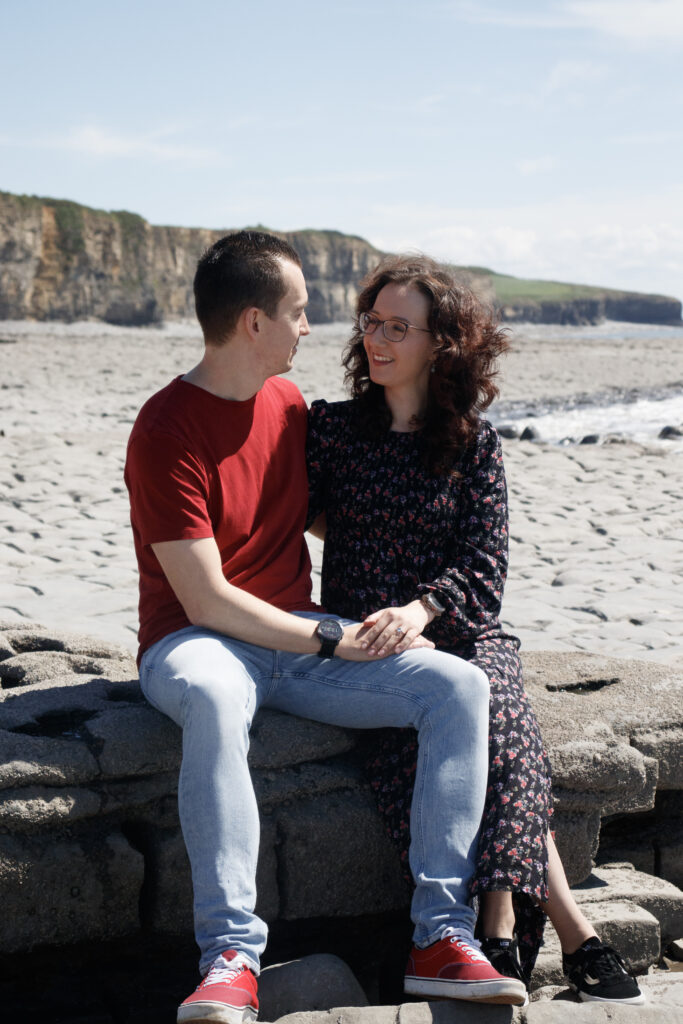 Couple sitting on a beach wall, smiling at each other with cliffs and ocean in the background.
