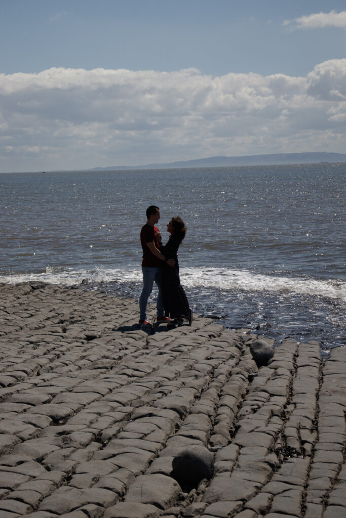 Couple embracing by the ocean on a sunny day, standing on rocky shoreline with scenic clouded sky.