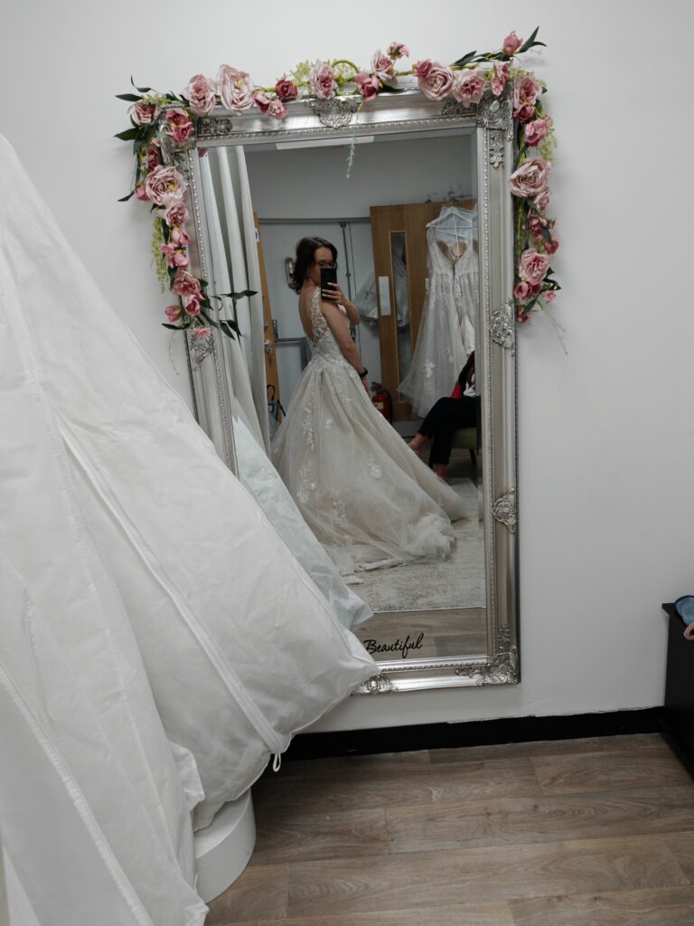 Bride trying on floral lace wedding dress in ornate mirror at bridal shop fitting room.